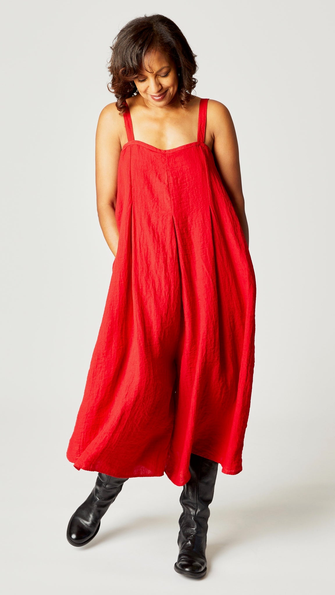 Model wearing red rayon jumpsuit with button shoulder straps, pleat details, and wide cropped leg with black leather boots.