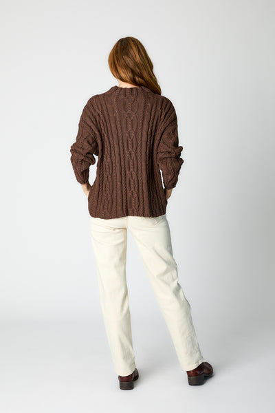 Rearview of model wearing chocolate cotton cable-knit sweater, ecru straight leg jeans, and brown leather boots. 