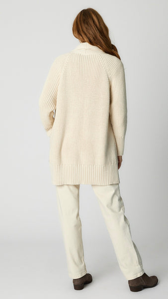 Rear view of model wearing cream cotton handknit oversized cardigan (without buttons) over natural bias top, ecru straight leg denim pant, and taupe suede boot. 