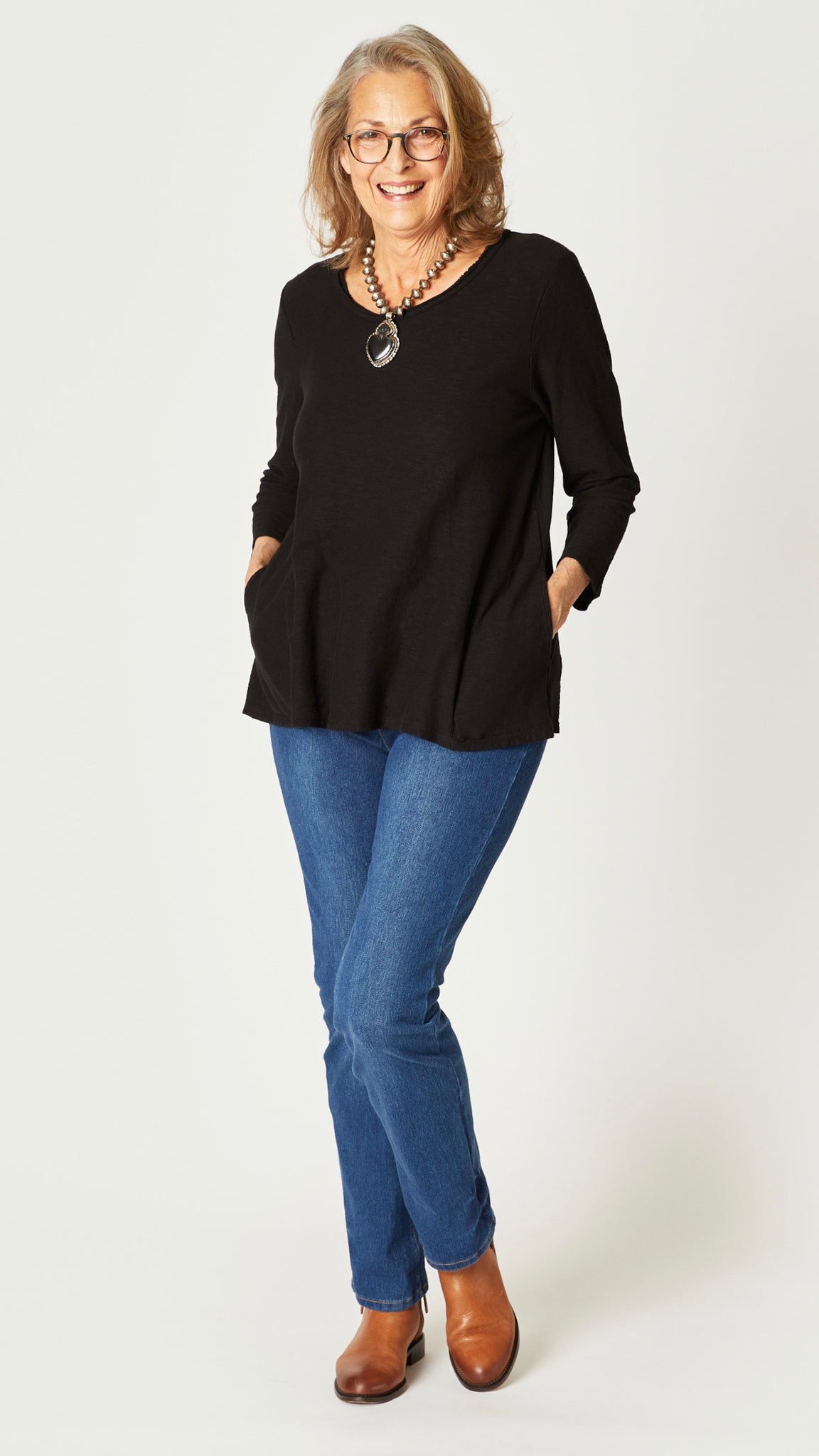 Model wearing black cotton-linen jersey top with 3/4 sleeves, hidden side pockets, and unfinished neckline, midwash straight leg jeans, and tan ankle boots.