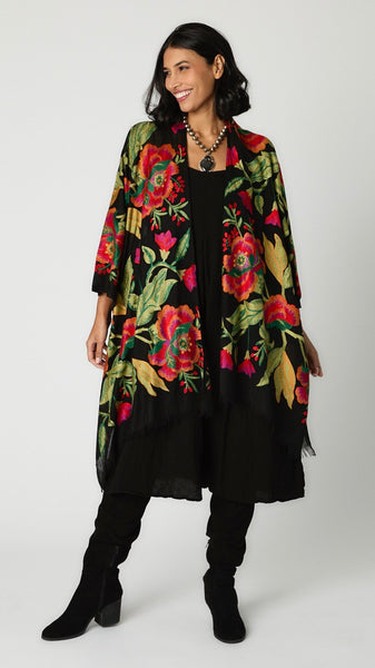 Model wearing black embroidered wool ruana with vibrant red and green florals, black rayon romper, and tall black suede boots.