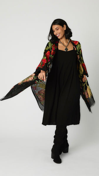 Model wearing black embroidered wool ruana with vibrant red and green florals, black rayon romper, and tall black suede boots.