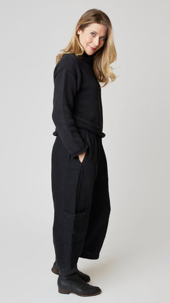 Model wearing black handknit cotton turtleneck sweater (slightly cropped), with black heavyweight linen cropped pant with side pockets above ankle darts, and black leather boots.