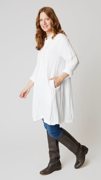 Model wearing white double cotton long sleeve tunic with standing collar, button-up placket and godet panels, with midwash denim leggings, and brown leather boots.