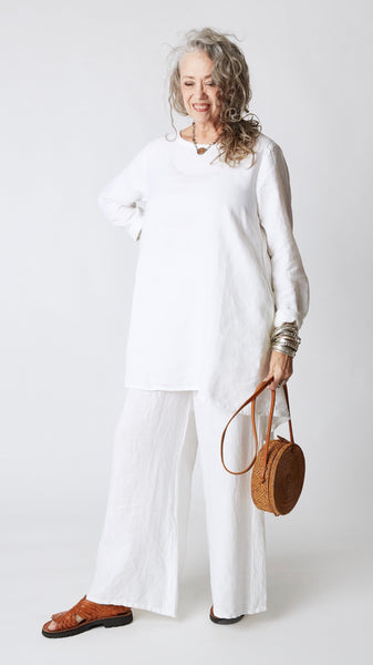 Model wearing white high-low linen tunic with long sleeves, wide-leg linen pants, silver bangles, round woven handbag, and brown leather sandals.