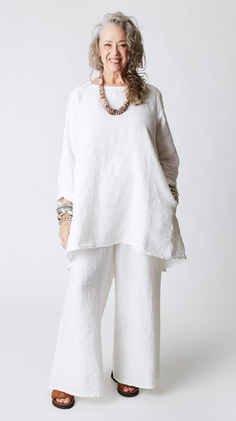 Model wearing white high-low linen tunic with long sleeves, wide-leg linen pants, silver bangles, coral disk necklace, and brown leather sandals.