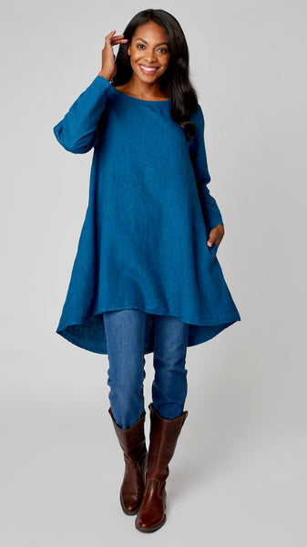 Model wearing deep sea high-low linen tunic with long sleeves, midwash denim leggings, and brown leather boots.