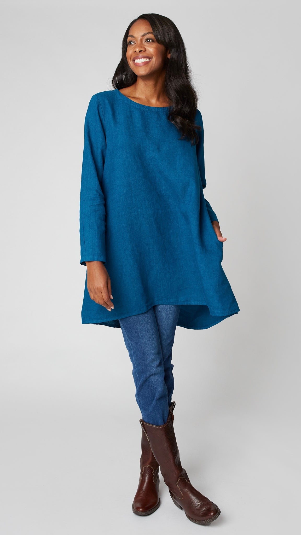 Model wearing deep sea high-low linen tunic with long sleeves, midwash denim leggings, and brown leather boots.