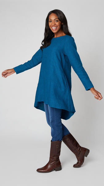 Model wearing deep sea high-low linen tunic with long sleeves, midwash denim leggings, and brown leather boots.