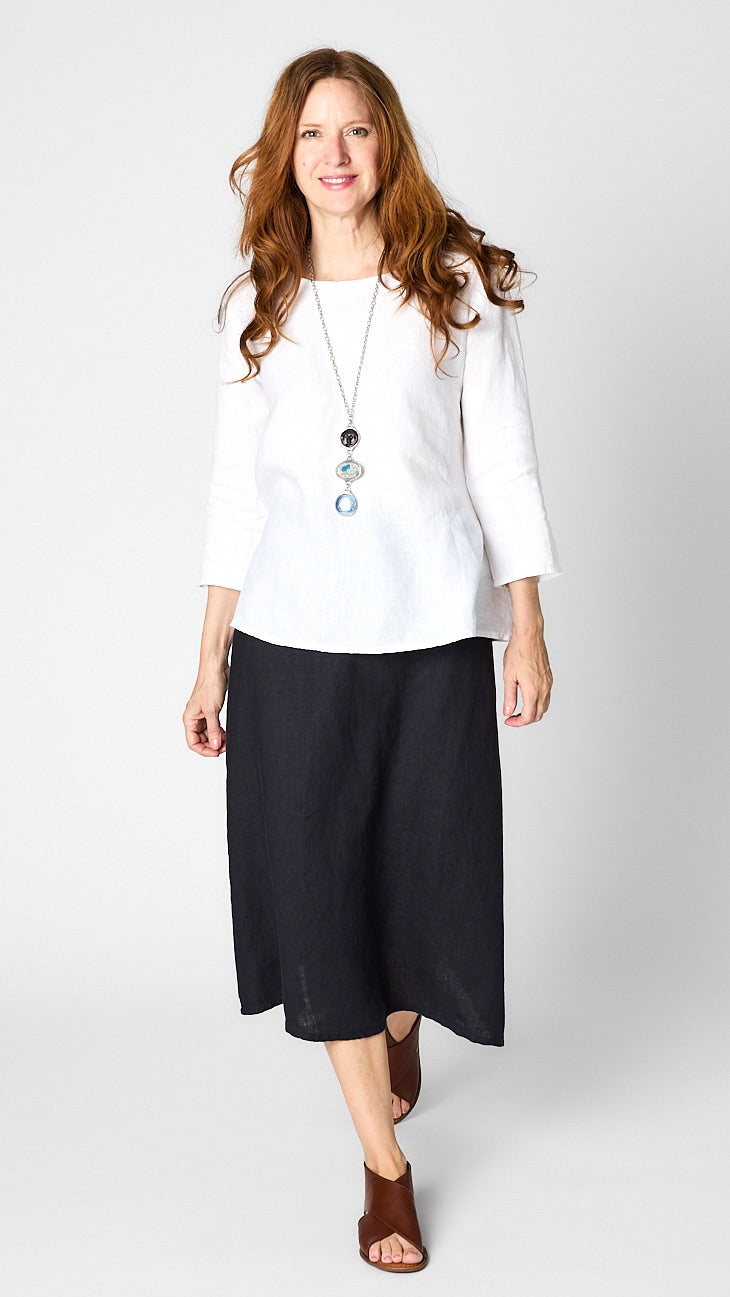 Model wearing white linen 3/4 sleeve shirt with A-line silhouette, ink A-Line Linen skirt, triple stone pendant necklace, and leather sandals. 