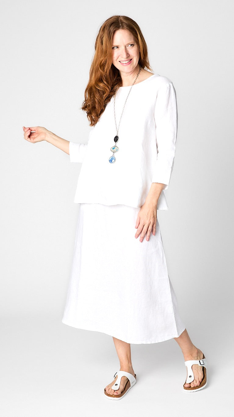 Model wearing white linen 3/4 sleeve shirt with A-line silhouette, white A-Line Linen skirt, triple stone pendant necklace, and leather sandals. 