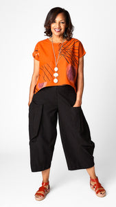 Model wearing orange batik printed short sleeve top with curved cropped hemline over black cropped double pocket pant in tencel cotton. 