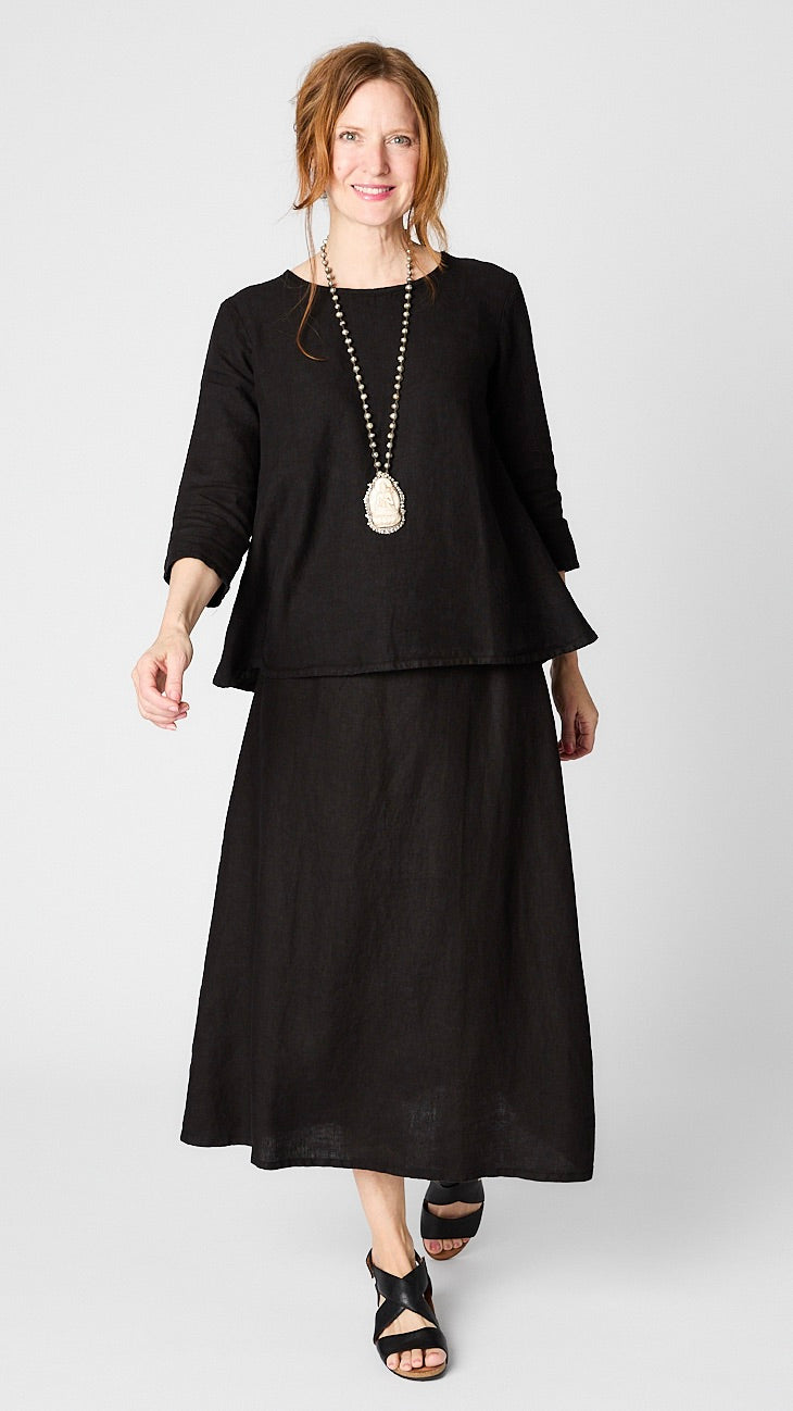 Model wearing black linen 3/4 sleeve shirt with A-line silhouette, black A-Line Linen skirt, bone pendant necklace, and leather sandals. 