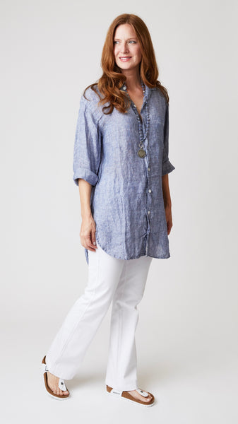 Model wearing chambray linen button-up tunic top with front pocket and shirttail hem, with white bootcut jeans and white sandals.