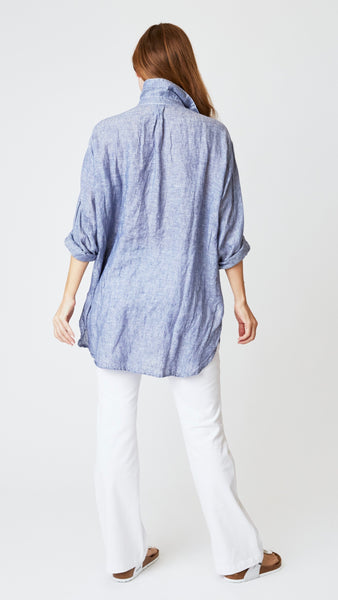 Rearview of model wearing chambray tunic with shirttail hem, displays box pleat detail at base of collar.  