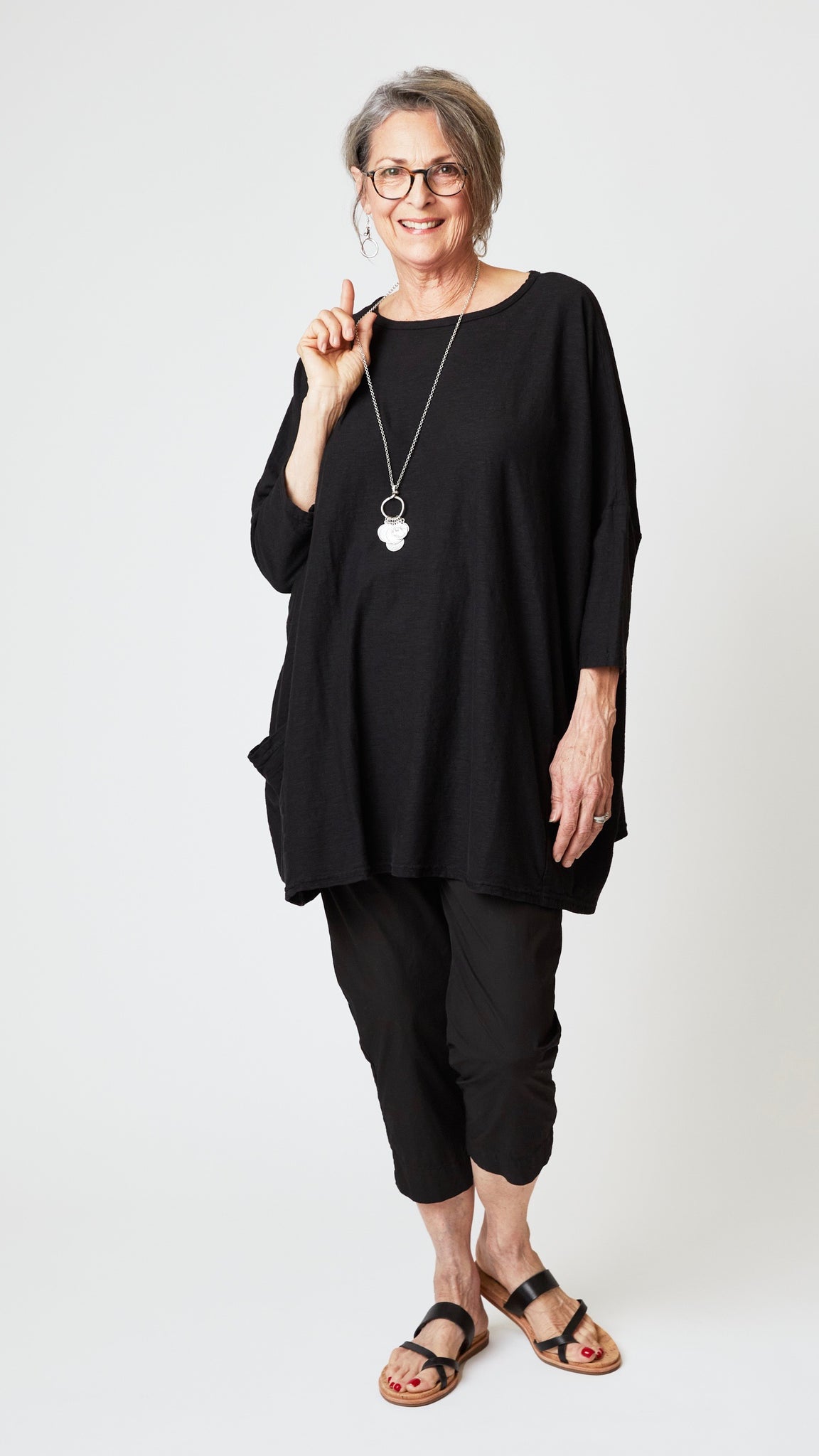 Model wearing oversized black cotton-linen jersey tunic with black cropped, tapered poplin cotton pant and black leather sandals. 