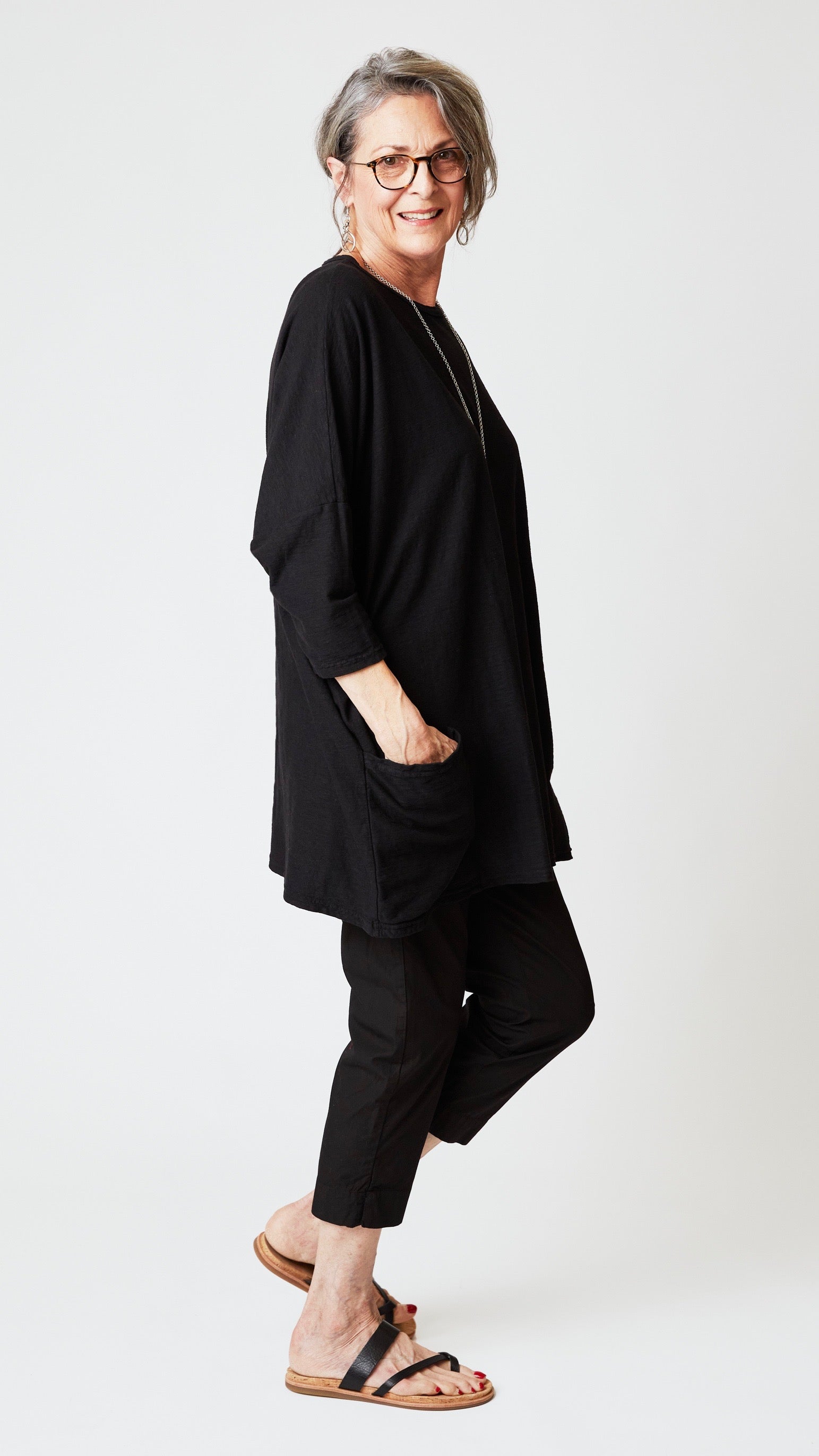 Model wearing black one size roomy two pocket tunic top, black tapered cropped pant in organic poplin, and black sandals. 