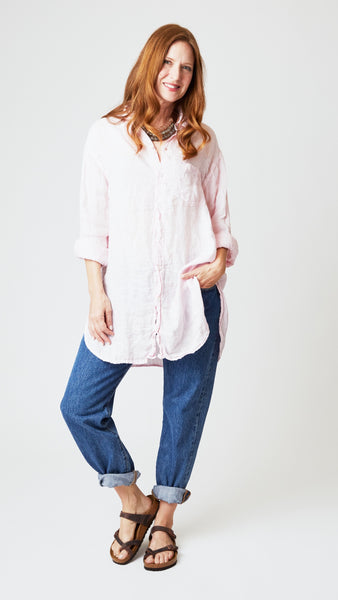 Model wearing pink chambray linen button up tunic with shirttail hem, front pocket, and midwash denim jeans, with beaded necklace and brown leather sandals. 
