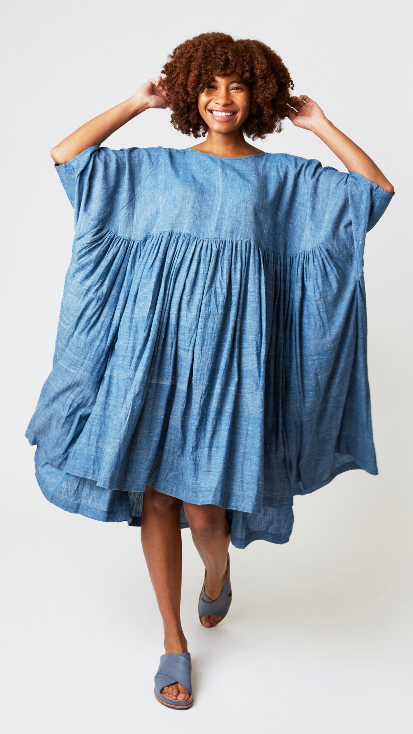 Model wearing oversized chambray blue cotton dress with raglan sleeves and waist-gathered skirt, and blue-gray sandals.