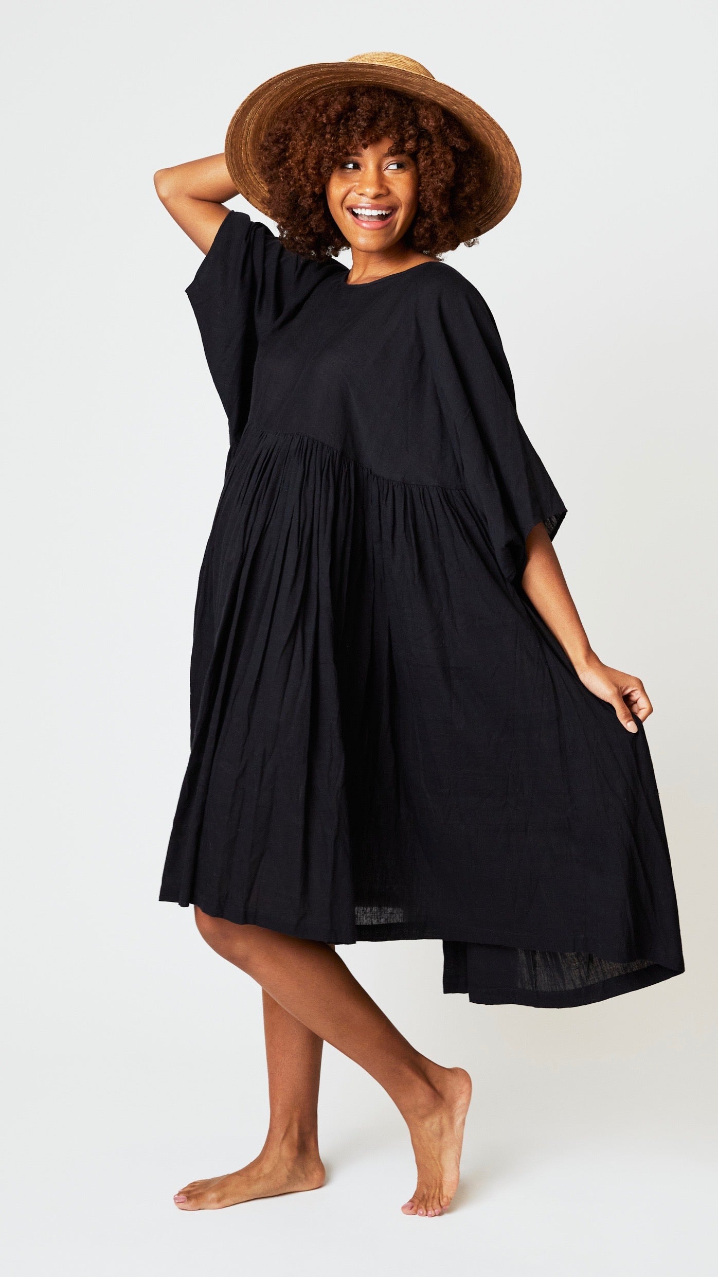 Model wearing oversized black cotton dress with raglan sleeves and waist-gathered skirt, and wide brimmed straw hat.