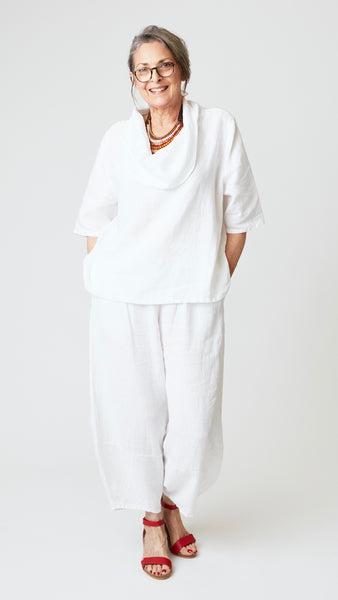 Model wearing white linen ankle length lantern pant, white linen cowl neck top with 3/4 sleeves, red beaded leather necklace, and red leather sandals. 