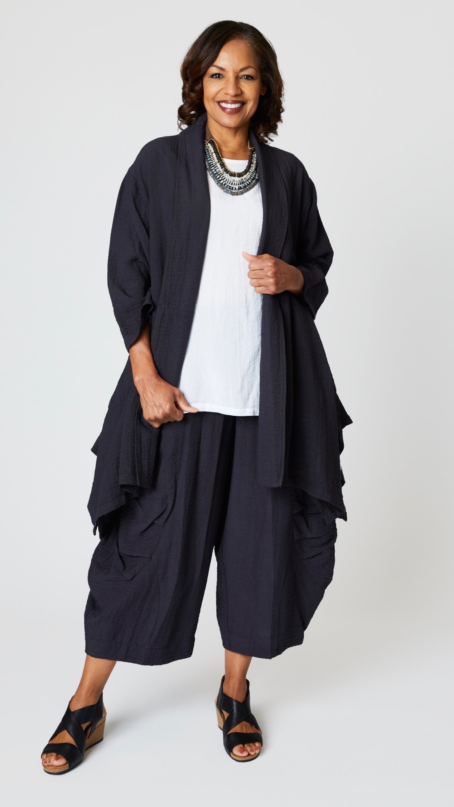 Model wearing "black pearl" rayon jacket with shawl collar, 3/4 sleeves, and tunic length hemline over white long tank,  cropped "black pearl" double pocket rayon pant, and black leather sandals. 