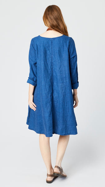 Rearview of model wearing indigo twill dress with v-neck, rolled long sleeves, and full skirt shows rear vertical seam detail.