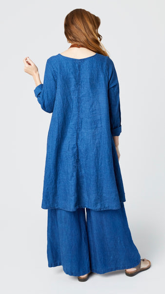 Rearview of model wearing indigo twill dress with v-neck, rolled long sleeves, and full skirt, and indigo twill wide-leg pants with brown leather sandals.