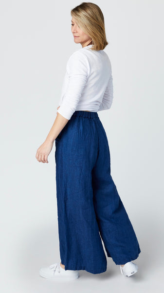 Model wearing high-waisted pants with wide-leg, flat front elastic waistband and side pockets, with white cotton/linen jersey bias cut top with 3/4 sleeves, and white sneakers.