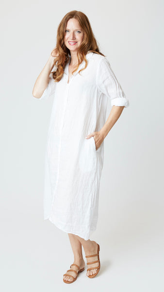 Model wearing white linen shirtdress with pointed collar and full length button-up panel, and tan leather sandals.