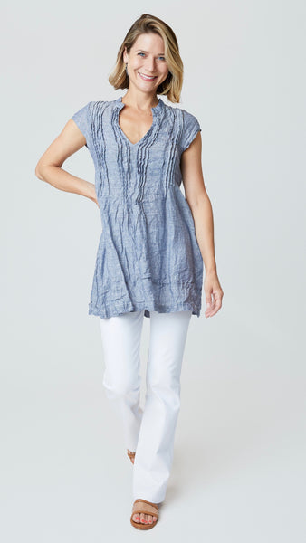 Model wearing chambray linen tunic with pin-tucked bodice, v-neck, and cap sleeves, with white bootcut denim, and tan leather sandals.