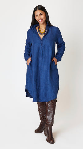 Model wearing indigo twill long sleeve dress with deep V-neck, collar, and shirttail hem, beaded leather necklace, and knee-high leather boots. 