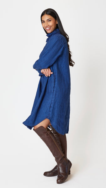 Model wearing indigo twill long sleeve dress with deep V-neck, collar, and shirttail hem, beaded leather necklace, and knee-high leather boots.