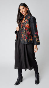 Model wearing shorter black silk jacket embroidered in shades of dusty pink, red, orange, and brown over a black rayon romper, black tights, and black leather ankle boots.