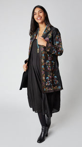 Model wearing black silk duster embroidered in dusty blue, purple, and brown tones over black rayon romper and black suede boots.