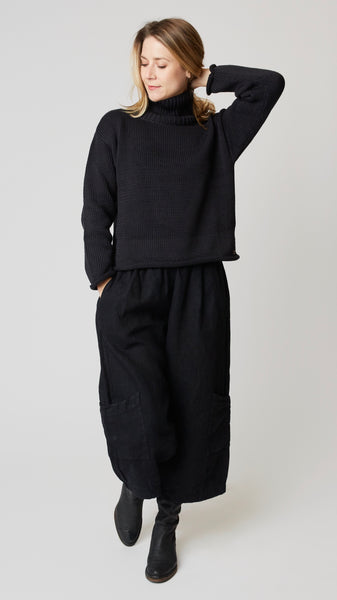 Model wearing black handknit cotton turtleneck sweater (slightly cropped), with black heavyweight linen cropped pant with side pockets above ankle darts, and black leather boots.