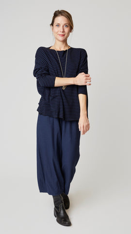 Model wearing navy jersey cropped dart pants, navy and black striped boxy long sleeve top, long silver chain with coin pendant, and black leather boots. 