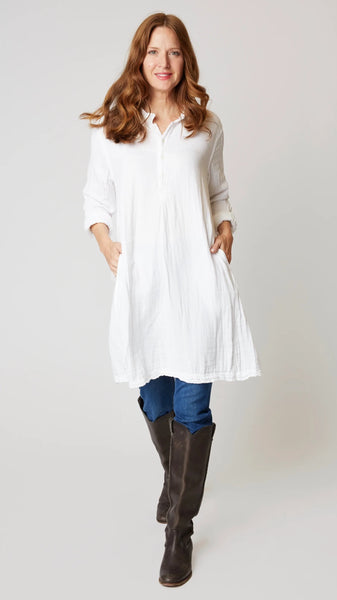 Model wearing white double cotton long sleeve tunic with standing collar, button-up placket and godet panels, with midwash denim leggings, and brown leather boots.