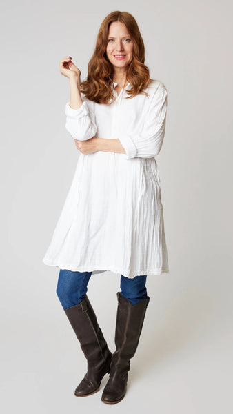 Model wearing white double cotton long sleeve tunic with standing collar, button-up placket and godet panels, with midwash denim leggings, and brown leather boots.