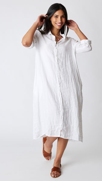 Model wearing white linen shirtdress with pointed collar and full length button-up panel, and brown leather sandals.