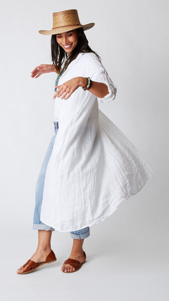 Model wearing white linen shirtdress as a duster with straw sunhat, turquoise necklace, white t-shirt, lightwash jeans, and leather sandals.