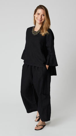 Model wearing black linen A-line top with scoop neck and 3/4 ruffle sleeves, black linen cropped pants, beaded leather necklace, and black flip-flops.