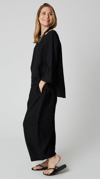 Model wearing black linen high-low A-line top with scoop neck and 3/4 ruffle sleeves, black linen cropped pants, beaded leather necklace, and black flip-flops.