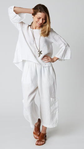 Model wearing white linen cropped pant with side pockets, white high-low top with 3/4 ruffle sleeve, and brown leather sandals.
