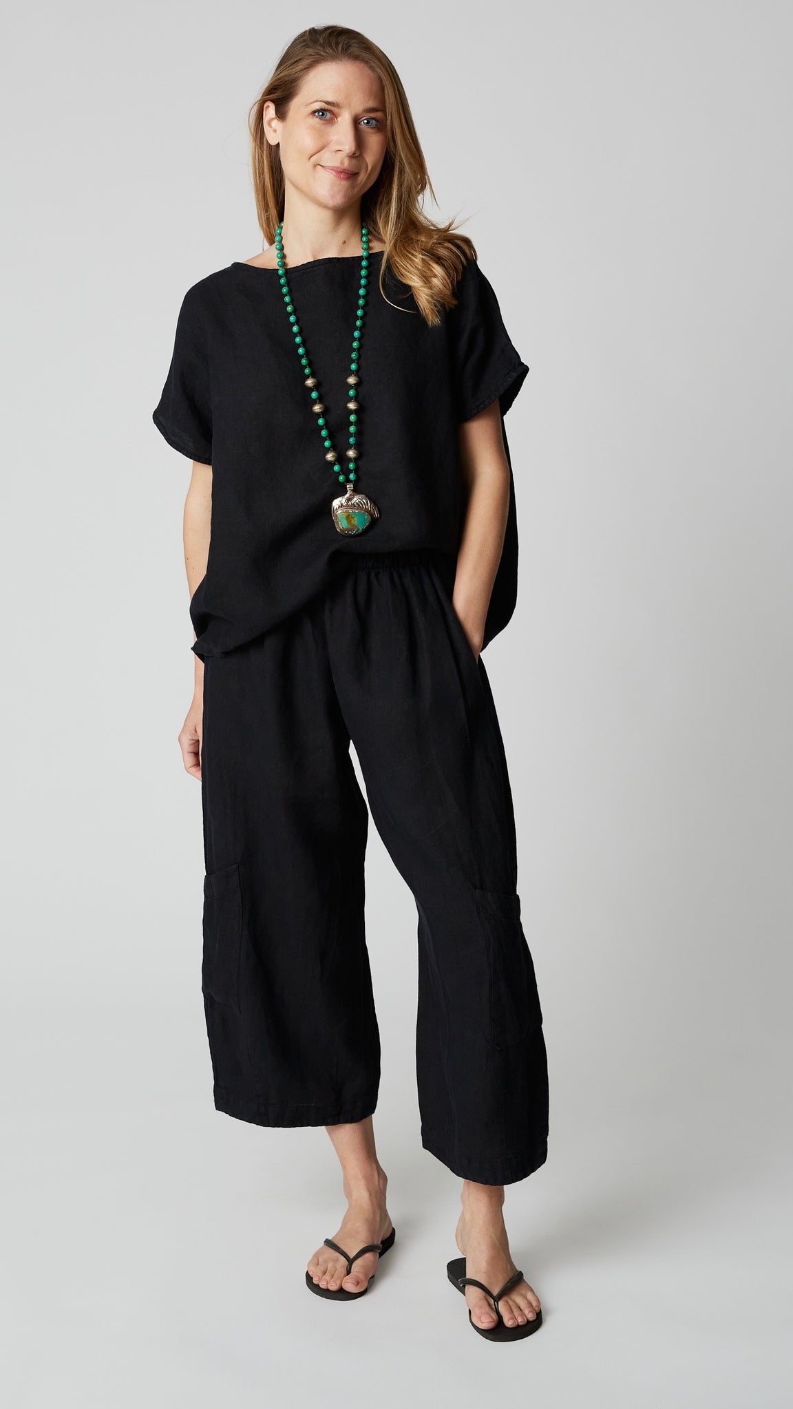 Model wearing black boxy cap-sleeve top with scoop neck, black linen cropped pant , turquoise necklace, and black flip-flops.